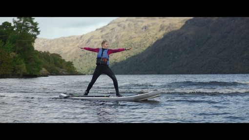 A woman stands on a paddleboard on a loch with mountains behind
