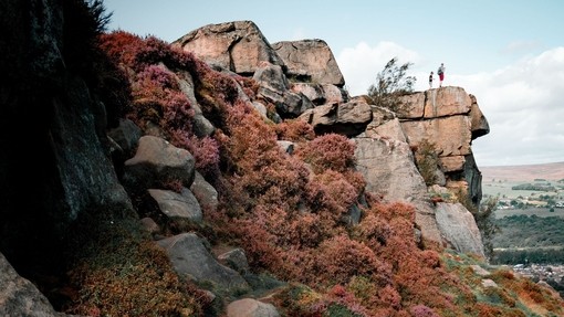 Two people in the distance stood on the edge of a rock looking out to the valley beyond, heather in foreground