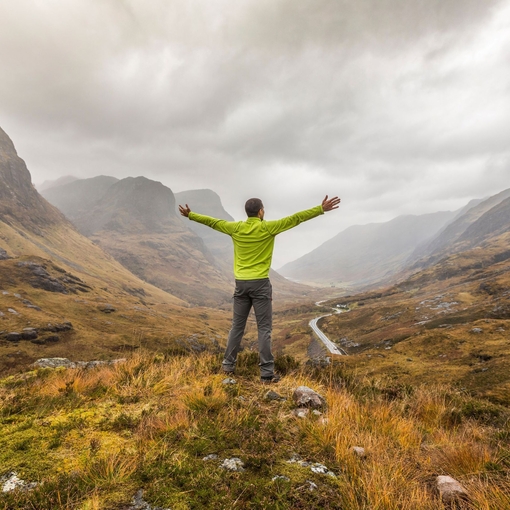 Back view of a man with arms outstretched standing in a valley looking at mountains cloaked in mist