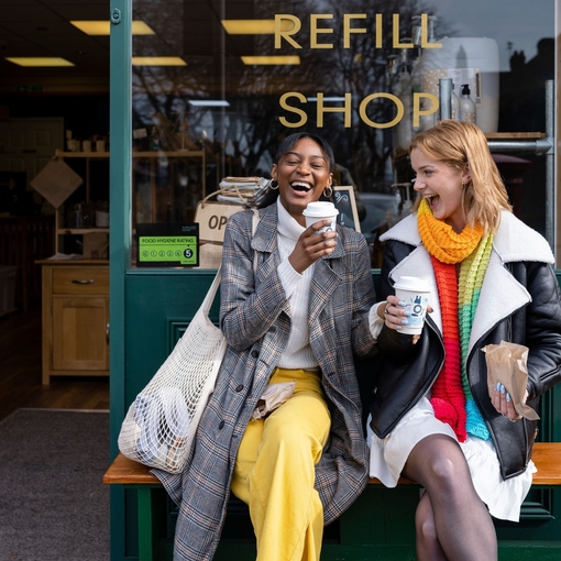 Two young women sat on a bench outside a coffee shop with takeaway coffees