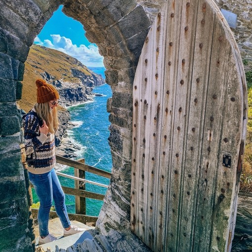 A woman stands at the top steps of a doorway at Tintagel with stunning coastline view beyond