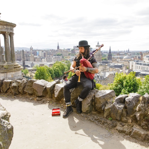 Man playing the bagpipes sat on a rock on a hill overlooking a city