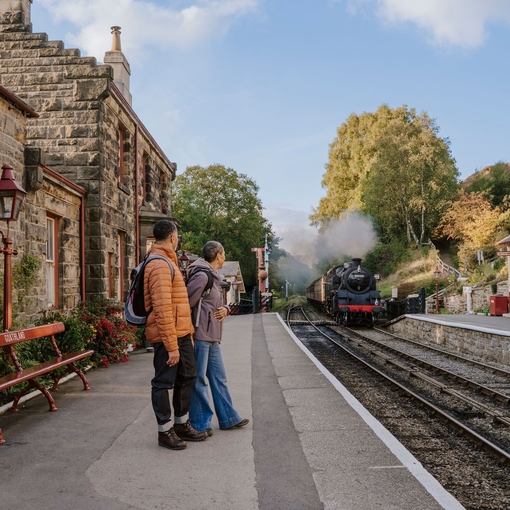 A man and woman standing on a train station platform as a train approaches in the background