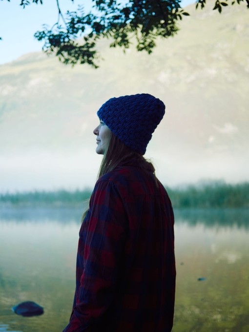Woman in hat standing in front of lake with mist and mountain backdrop.