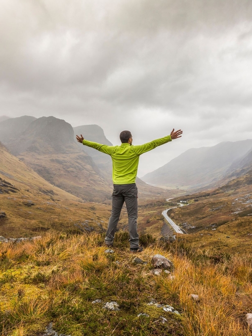 Back view of a man with arms outstretched standing in a valley looking at mountains cloaked in mist 