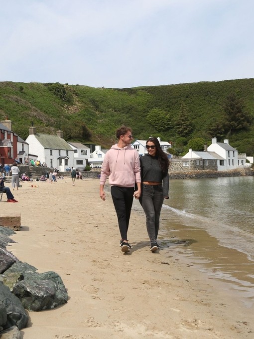 Two people walking along a sandy beach with the ocean to one side and pubs and shops on the other side.