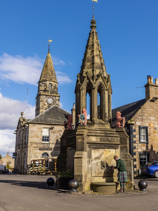 A man gets water from a large stone fountain with faded red lion statues in a town square.
