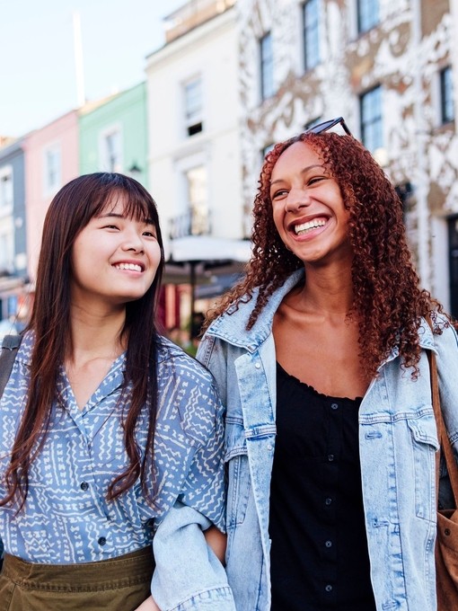 Two young women linking arms and walking in a street smiling