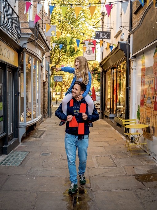 A man and his child on his shoulders walk through a quaint village shopping street.