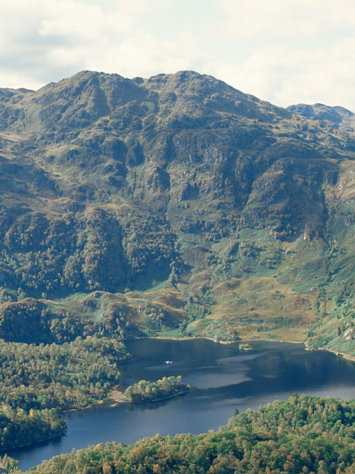 A distant view of a loch, surrounded by trees and green mountains.