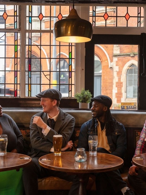 Four friends smiling and seated with drink in a old style pub with antique stained glass window behind them