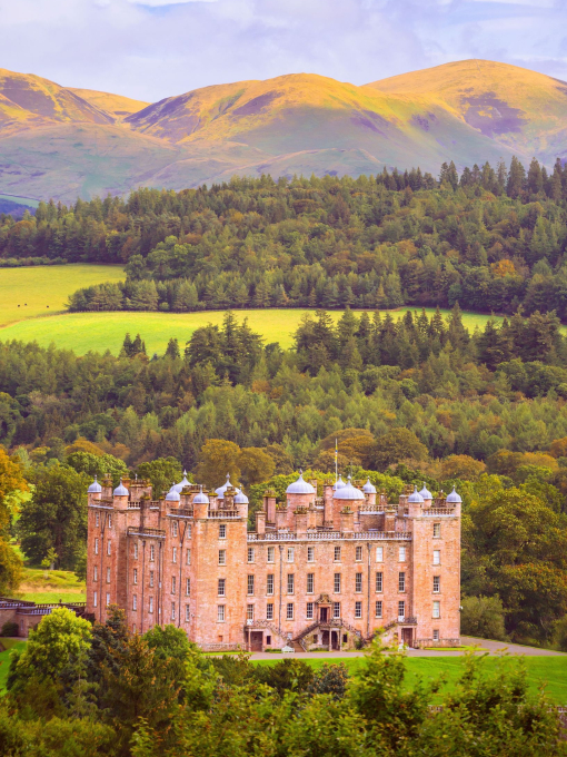 Exterior of a sandstone palace with ornate turrets surrounded by woodland with high mountains in the distance in autumn.
