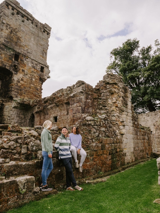 Two women and a man sitting on the ruins of a large castle and garden area.