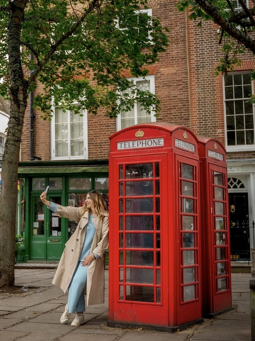 A woman leans against a red telephone box and takes a selfie in a town square.