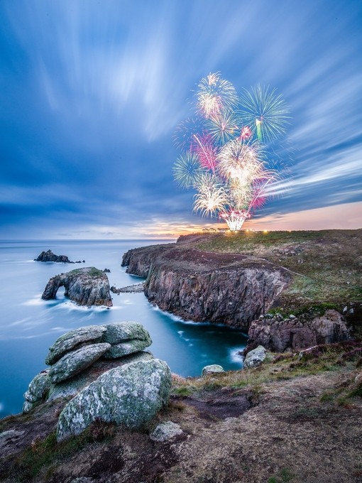 Firework display seen from across a rocky headland at twilight