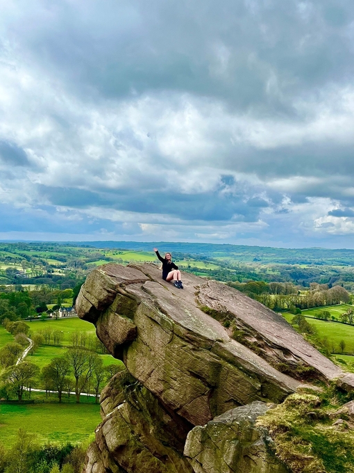 A seated woman waving from a hanging stone, overlooking a country panorama.