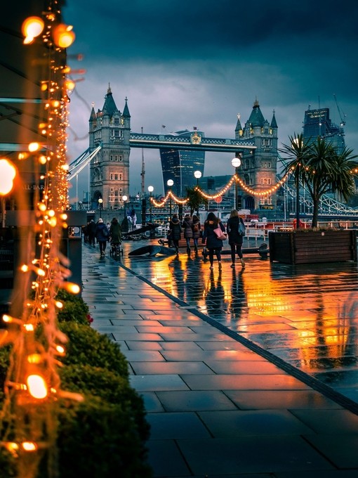 Fußgänger, die an einem verregneten Abend an Lichterketten vorbeigehen, im Hintergrund die Tower Bridge, London