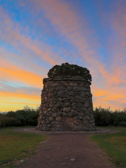A small stone tower with a plaque on it surrounded by grass and moor under a vibrant blue and orange sky at sunset.