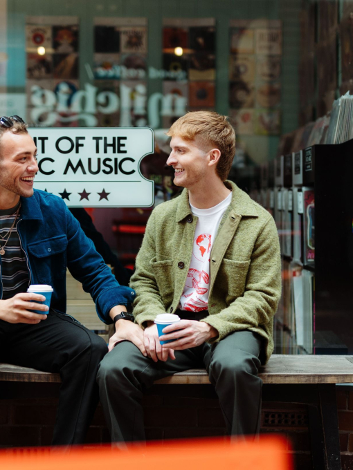 Two men sit with take away drinks in front of a shop