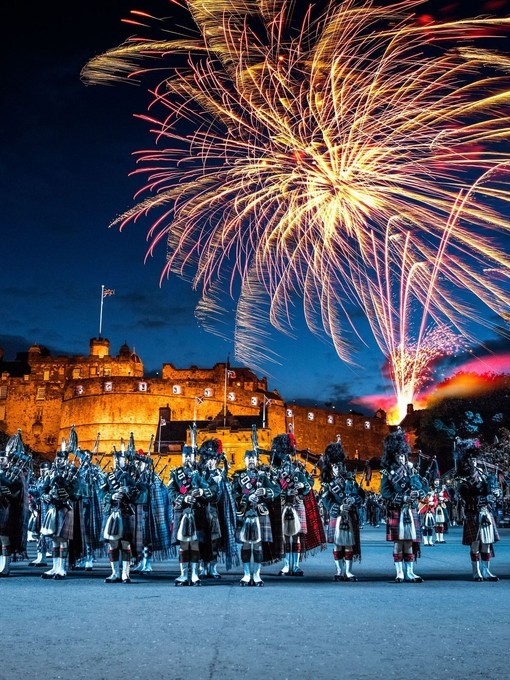 The arena at the Edinburgh Military Tattoo during a performance of the military event, parade ground and packed spectator seating. A light show projecting onto the castle walls. Marching band with a leading conductor, and massed pipers playing the bagpipes. Fireworks exploding in the night sky.