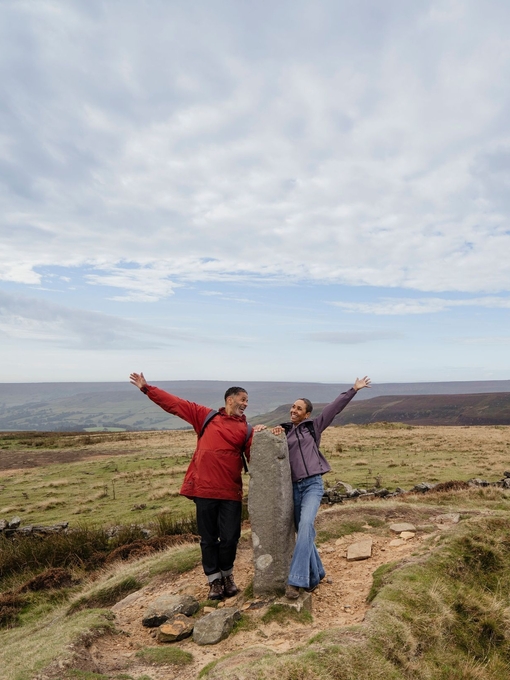 A man and woman standing around a large stone with arms outstretched in a scenic countryside landscape