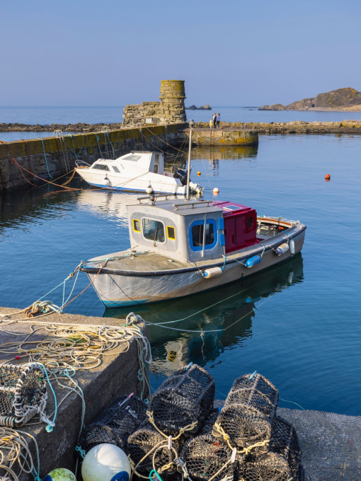 Two small boats moored in a small stone harbour on a sunny day, with seven lobster cages in the foreground.
