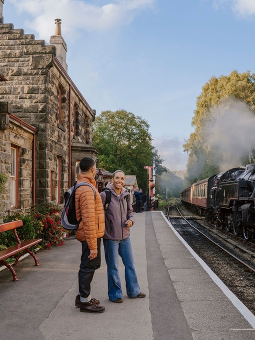 A man and woman standing on a train station platform as a train approaches in the background