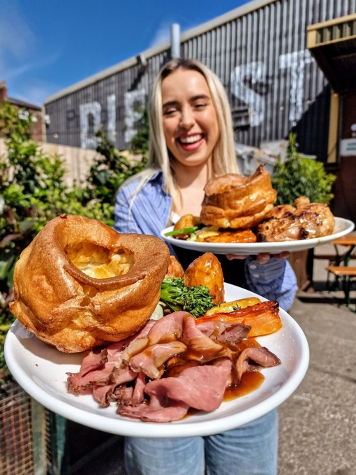 A smiling woman holding large plates of pub food in a large beer garden.