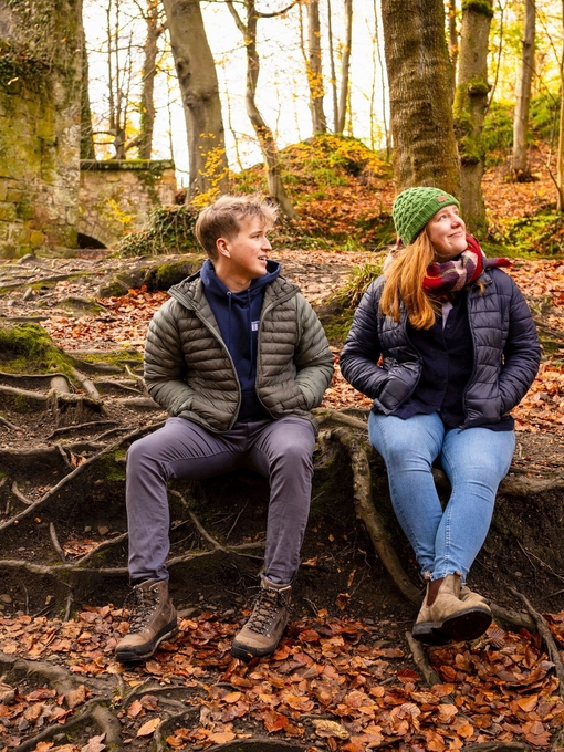 A man and woman in hiking gear smile at each other while sitting on tree roots in a forest.