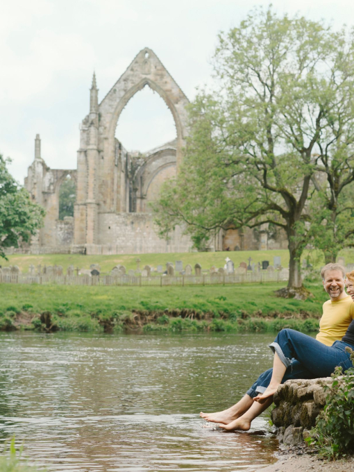 A man and a woman sit by a river near a heritage Abbey