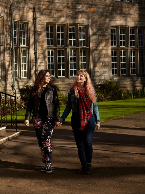 Two women holding hands whilst walking past a university building.