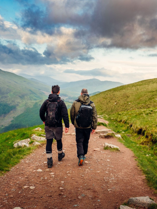 Two male hikers trekking along a pebbled path, cloudy skies, green rolling mountain vista.