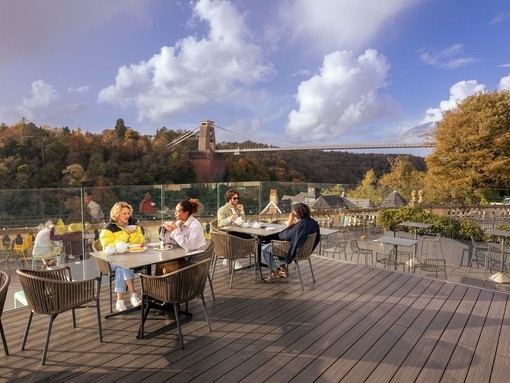 Friends enjoying tea and cake on a rooftop terrace, overlooking a river and suspension bridge.