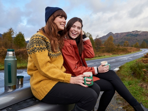 Zwei Frauen machen an einer Landstraße in den schottischen Highlands Zwischenpause und trinken Kaffee aus einer Thermoskanne.