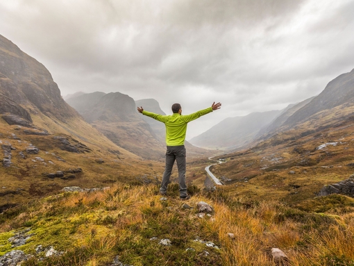 Back view of a man with arms outstretched standing in a valley looking at mountains cloaked in mist 