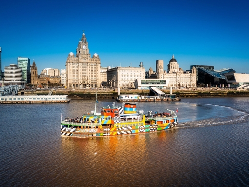 A colourful ferry crossing a river with a city backdrop and bright blue sky