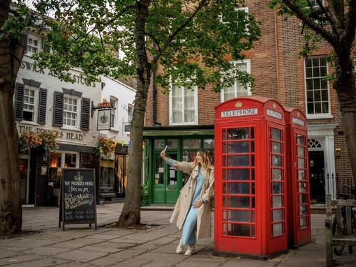 A woman leans against a red telephone box and takes a selfie in a town square.