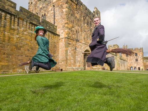 Two wizarding professors demonstrating flying on a broom outside a large castle.
