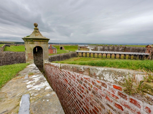A brick defensive wall with a small tower amid a larger stone fortification under grey skies.