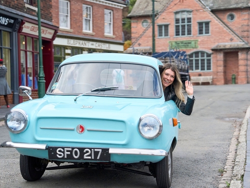 A woman waving and smiling inside a vintage car at a living museum