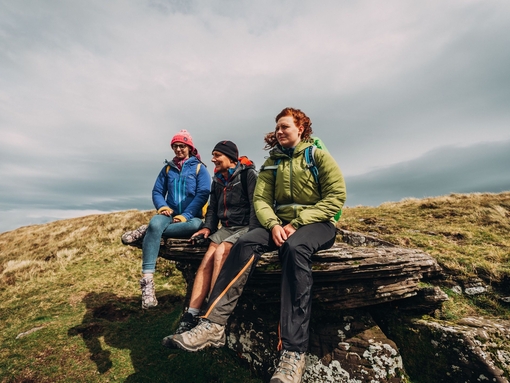 Three people sitting on a rock along a hiking route beneath a stormy sky.
