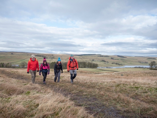 Four people hiking across a country hilltop path in the day with grey clouds in the sky.
