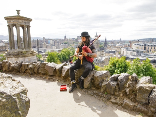 Man playing the bagpipes sat on a rock on a hill overlooking a city