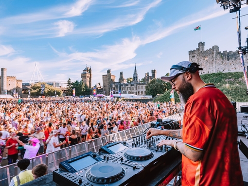 A DJ using music decks on a stage overlooking a crowd in the grounds of a castle