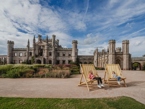 Man and woman holding ice creams in oversized deck chairs in manicured castle grounds and gardens.