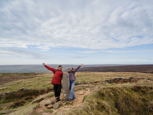 Un homme et une femme debout autour d'un grand rocher, les bras tendus, dans un paysage champêtre