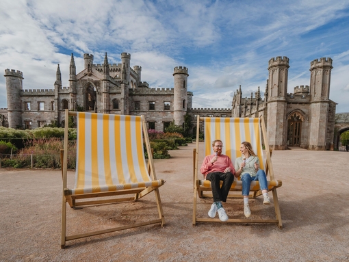 Man and woman holding ice creams in oversized deck chairs in manicured castle grounds and gardens.