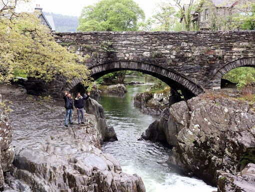 Two men waving from some rugged rocks beneath a stone bridge over a narrow river