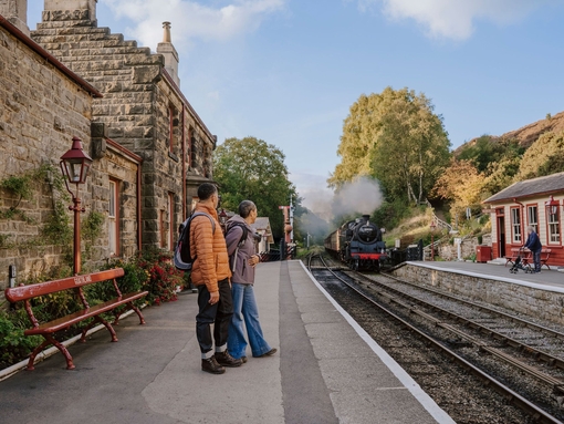 A man and woman standing on a train station platform as a train approaches in the background