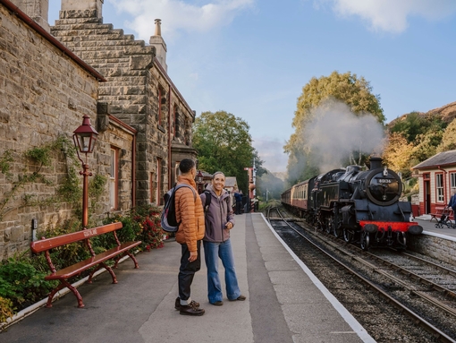 A man and woman standing on a train station platform as a train approaches in the background